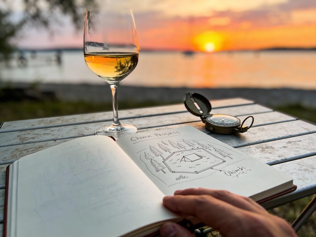 Work-in-Progress Shot: Ein Hand-Notizbuch liegt auf einem Campingtisch, darauf eine Skizze der Stellplatzanordnung. Daneben ein Kompass und ein Weinglas. Im Hintergrund verschwommen der Sonnenuntergang über dem Chiemsee.