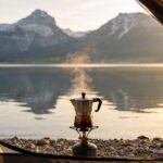 Ein halboffenes Zelt direkt am Kiesufer eines bayerischen Sees. Im Vordergrund ein Gaskocher mit dampfendem Kaffee, im Hintergrund unscharf die Alpenkette, die sich im glatten Wasser spiegelt. Morgenlicht.
