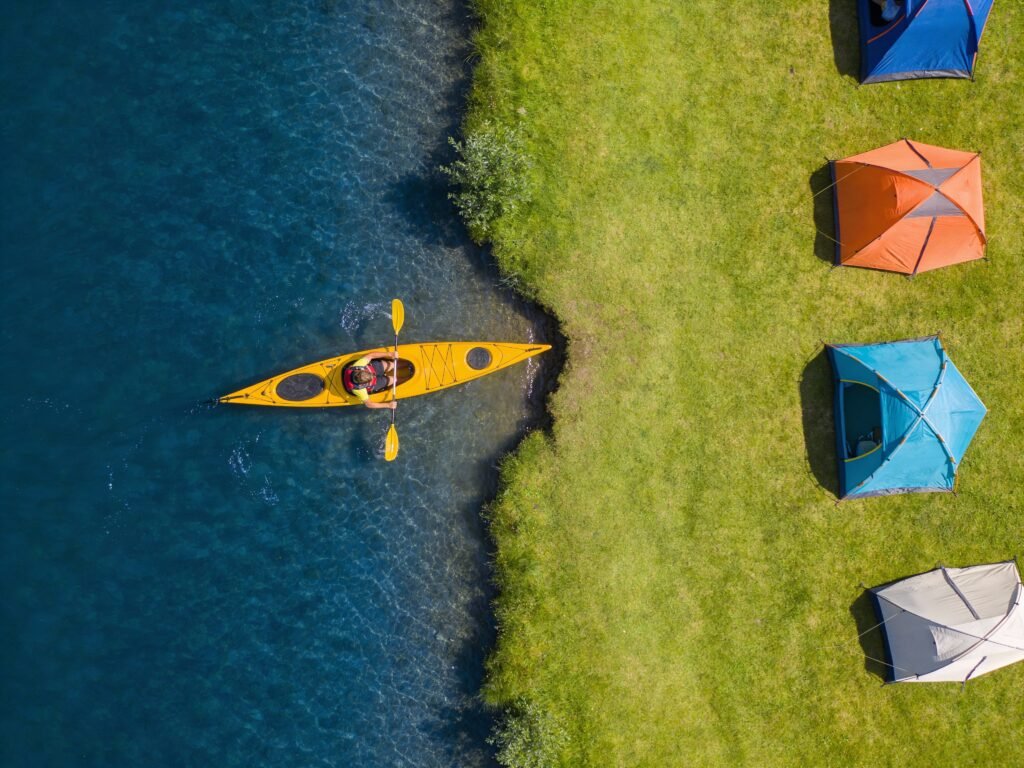Drohnenaufnahme (simuliert): Senkrechte Sicht nach unten. Links tiefblaues Wasser, rechts hellgrüne Wiese mit bunten Zelten. Ein gelbes Kajak wird gerade ins Wasser gelassen.